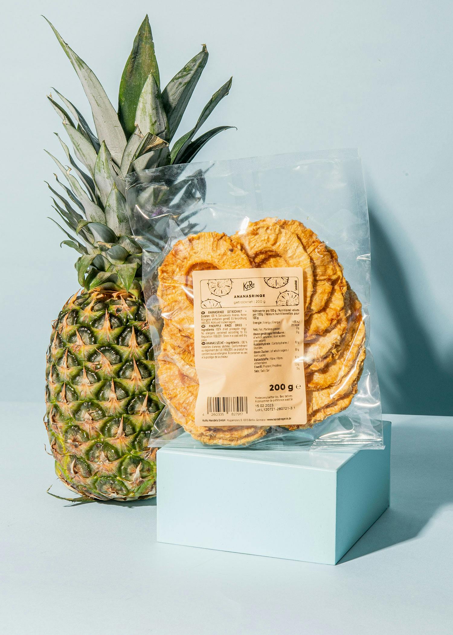 eine Ananas steht neben einem Plastikbeutel mit 200 g getrockneten Ananasscheiben