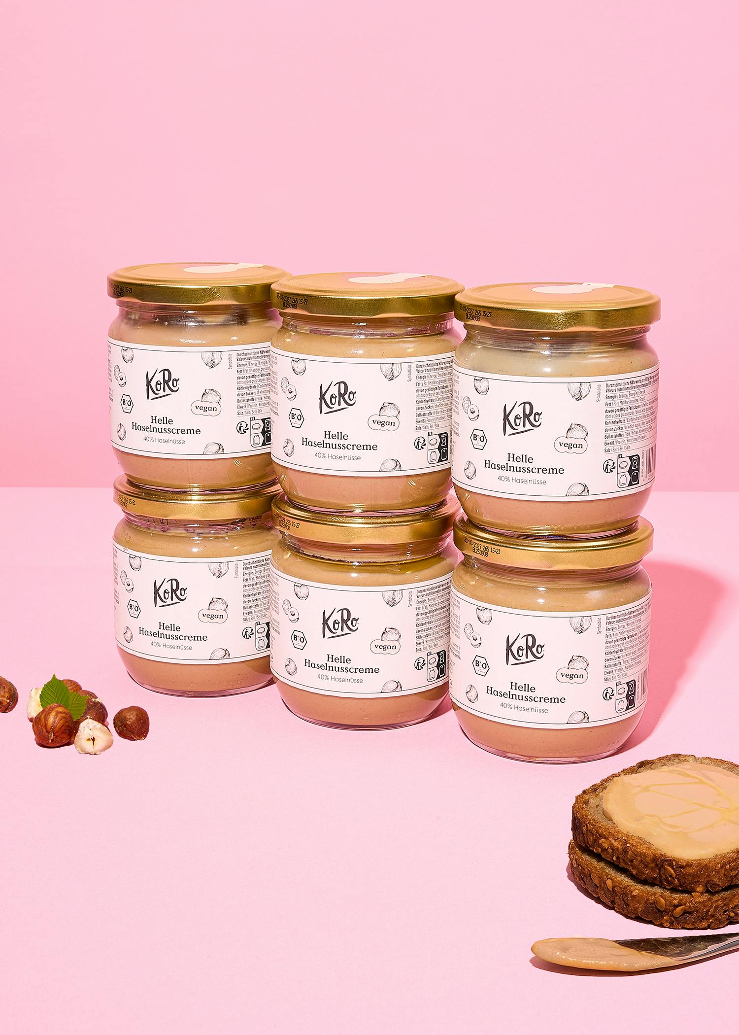 sechs gläser koro helle haselnusscreme sind auf einem rosa hintergrund gestapelt, daneben liegen einige haselnüsse und ein brot mit aufstrich.
