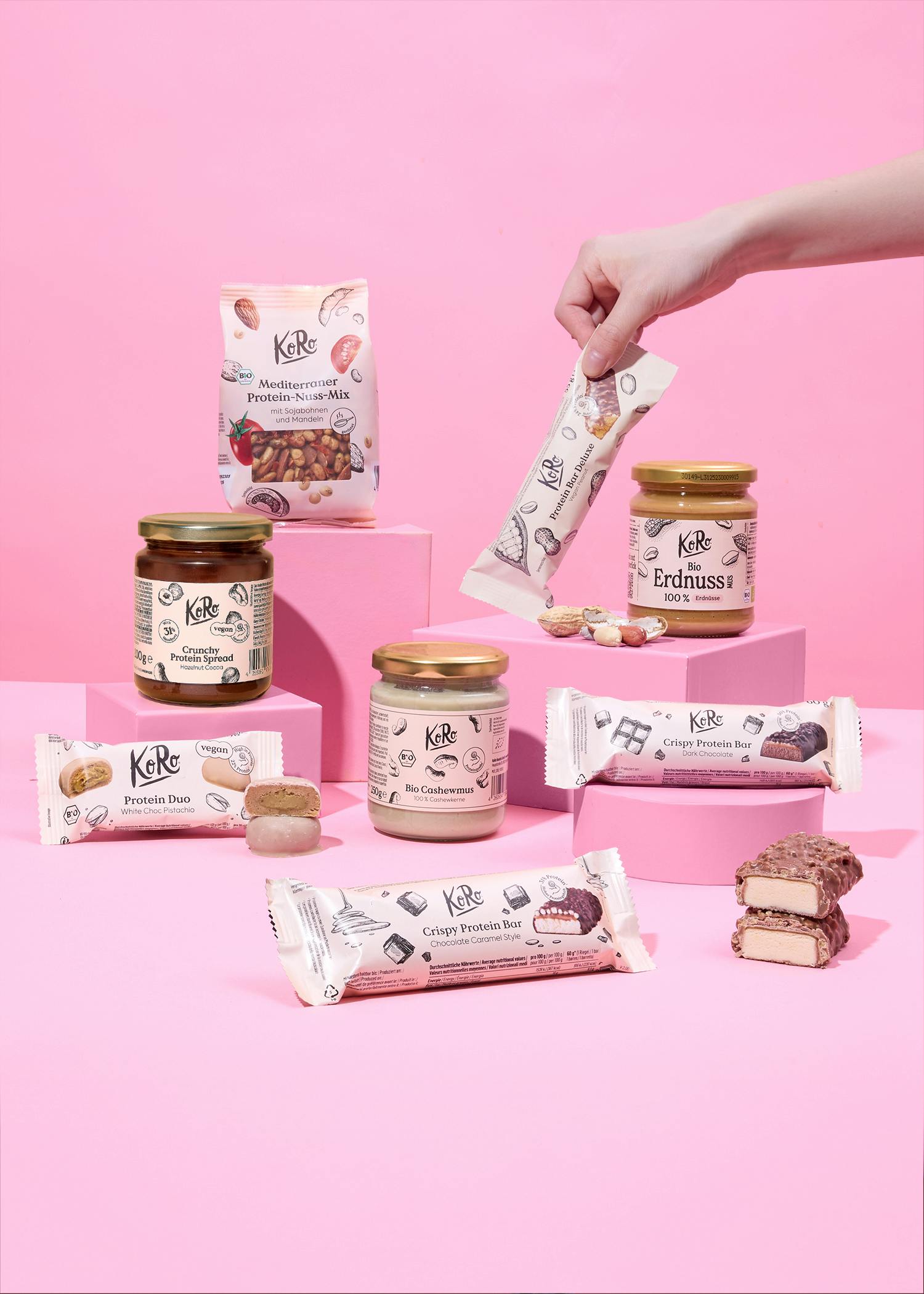 eine hand hält einen proteinriegel inmitten einer auswahl an koro-produkten wie nussmusen, proteinriegeln und einem nuss-mix, die auf rosa podesten vor einem rosa hintergrund präsentiert werden.