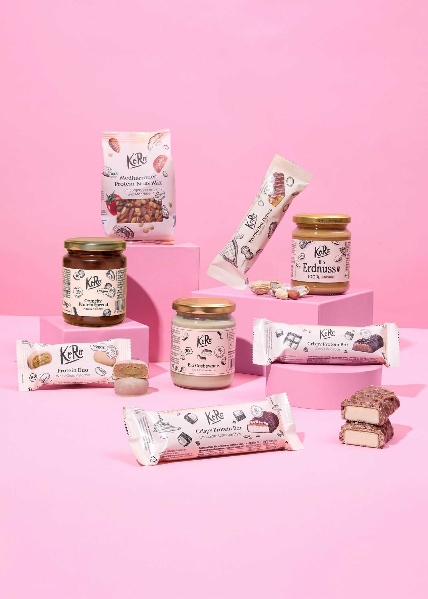 verschiedene koro-produkte wie proteinriegel, nussmuse und nussmischungen sind auf rosa blöcken vor einem rosa hintergrund arrangiert.