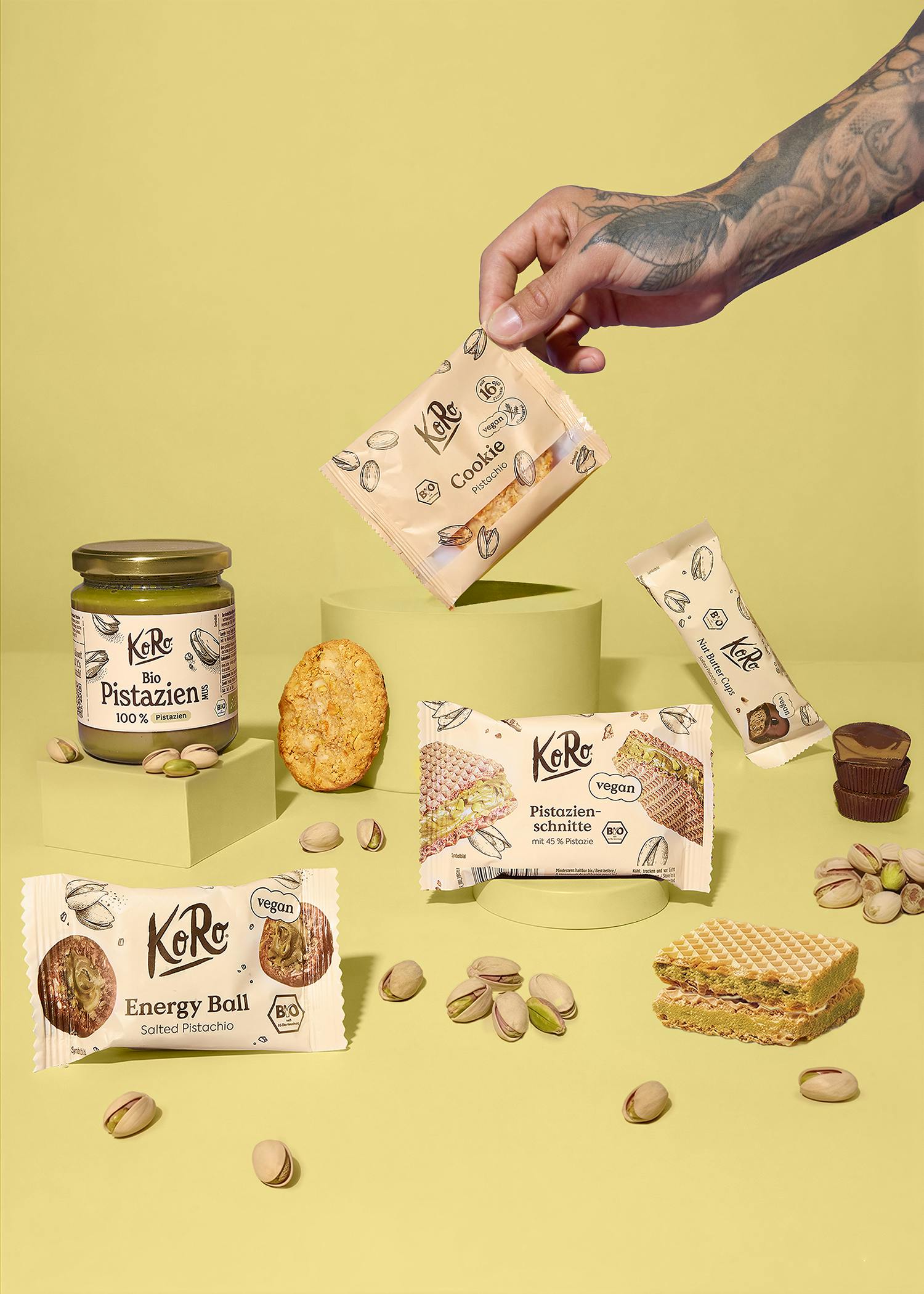 eine anordnung von verschiedenen koro-pistazienprodukten, darunter aufstriche, kekse, riegel und waffeln, zusammen mit losen pistazien auf einem gelben hintergrund, wobei eine hand einen keks hält.