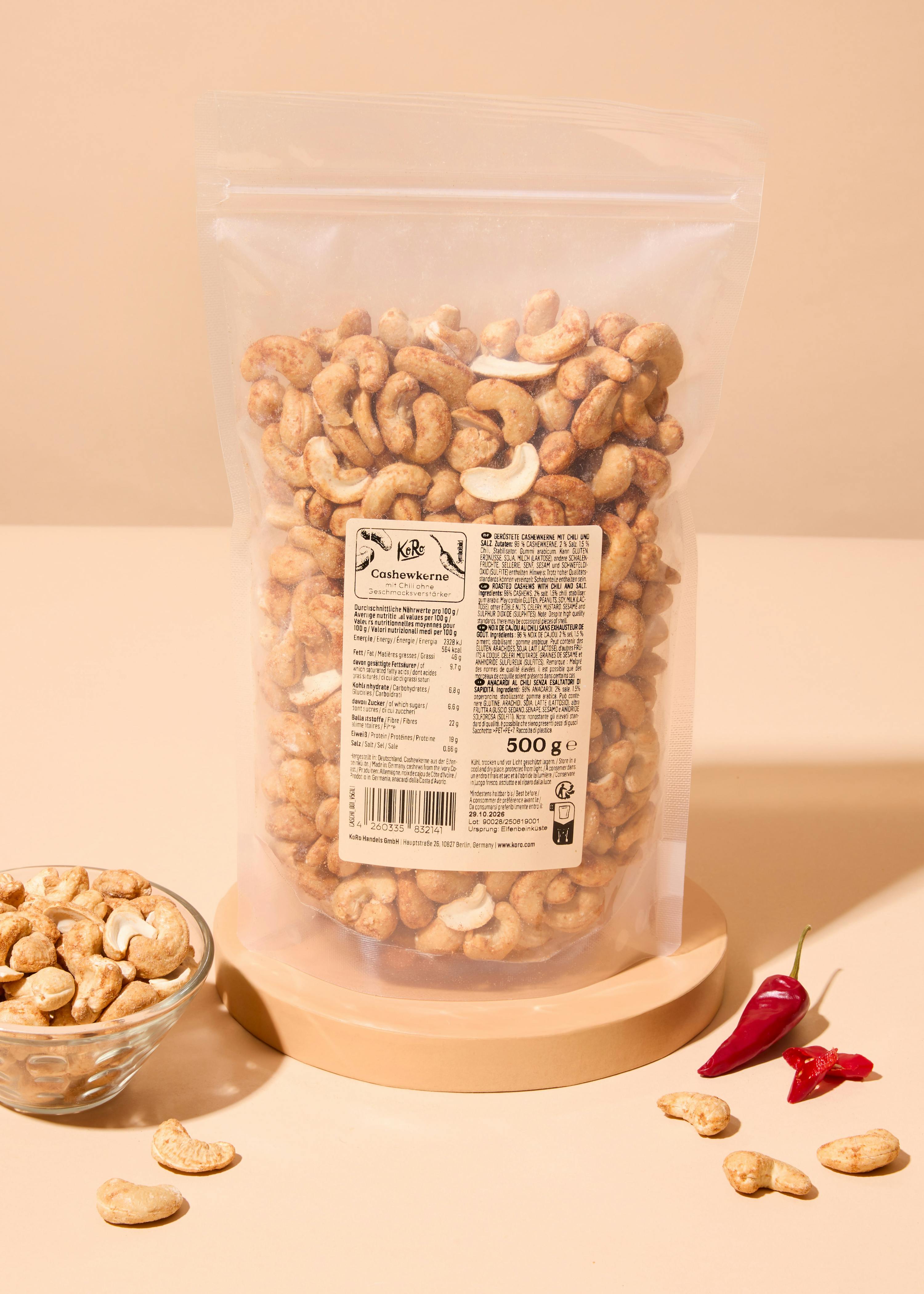 eine durchsichtige plastiktüte mit gerösteten cashewkernen mit chili und salz steht auf einer hellbraunen oberfläche, daneben eine kleine glasschale mit cashewkernen und zwei rote chilischoten.