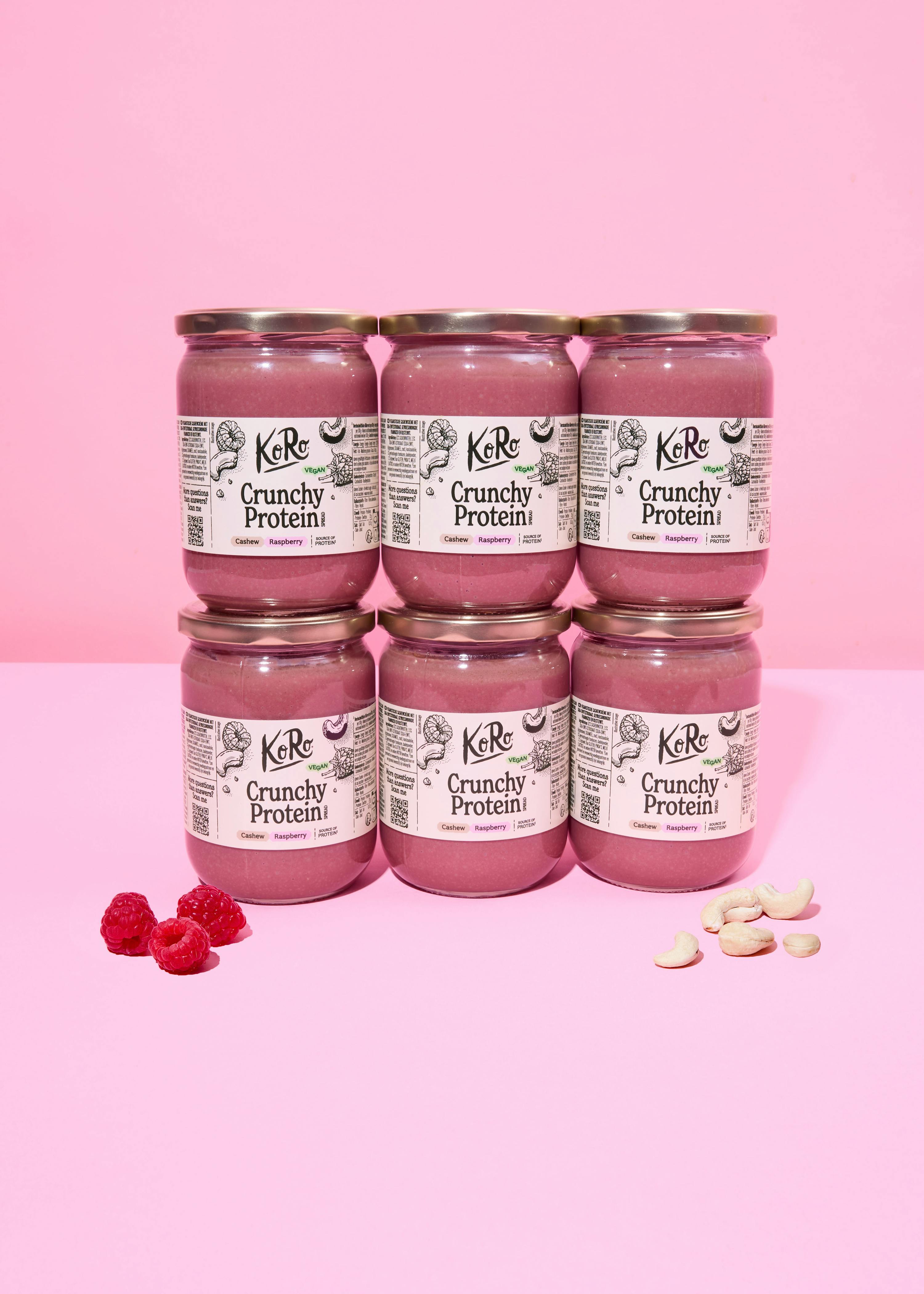 sechs gläser koro crunchy protein cashew-himbeer-aufstrich, in zwei reihen gestapelt auf einem rosa hintergrund, mit frischen himbeeren und cashewnüssen.