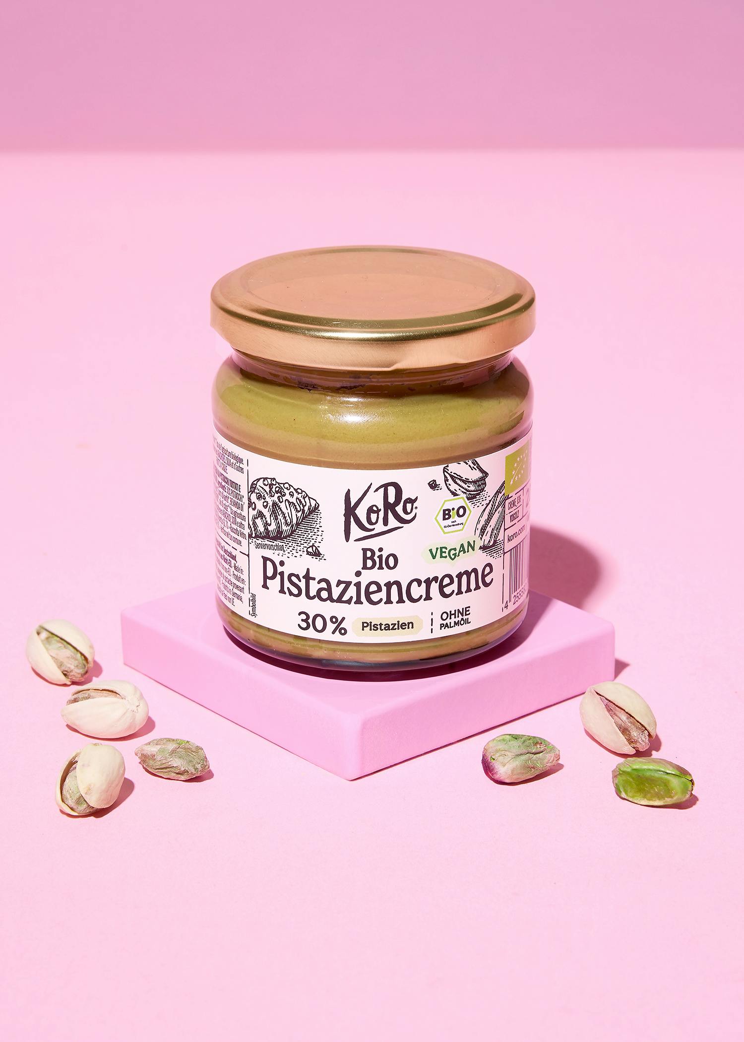 ein glas koro bio pistaziencreme steht auf einem rosa block, umgeben von mehreren pistazien.