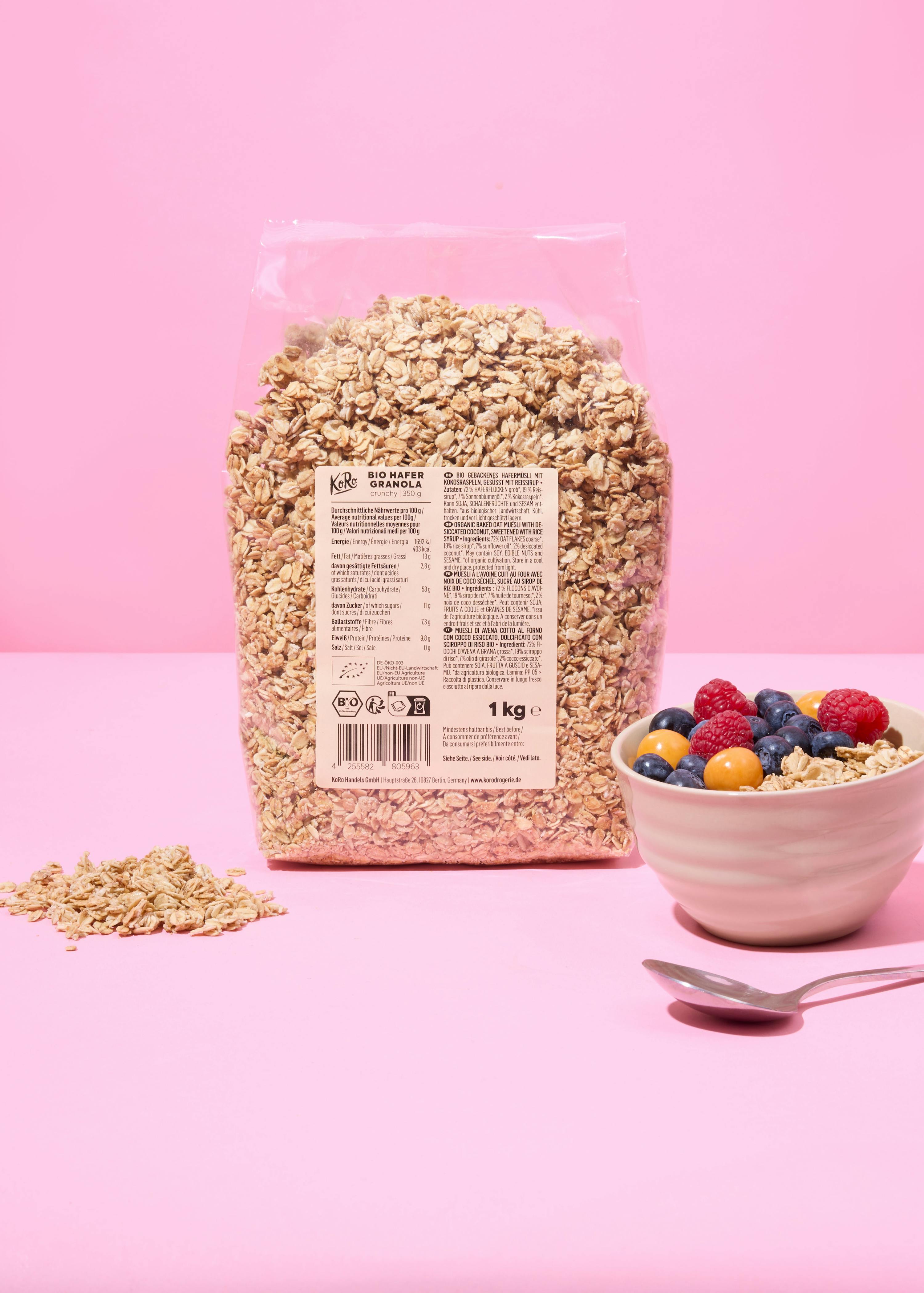 eine große transparente tüte mit bio-hafer-granola steht neben einer weißen schale mit granola, beeren und physalis sowie einem löffel auf einem pinkfarbenen hintergrund.