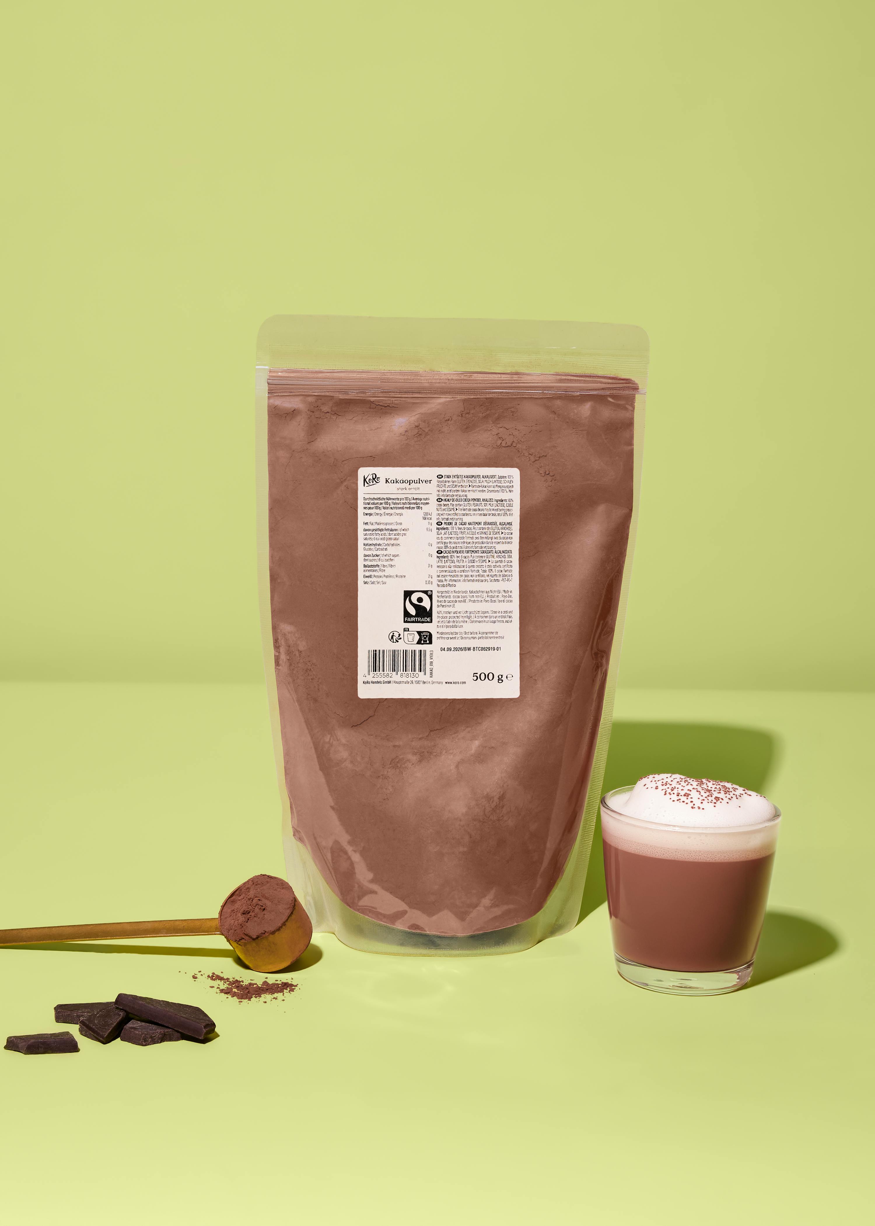 eine transparente verpackung mit kakaopulver, ein holzlöffel mit kakaopulver, schokoladenstücke und ein glas heißer kakao mit schaum auf einem hellgrünen hintergrund.