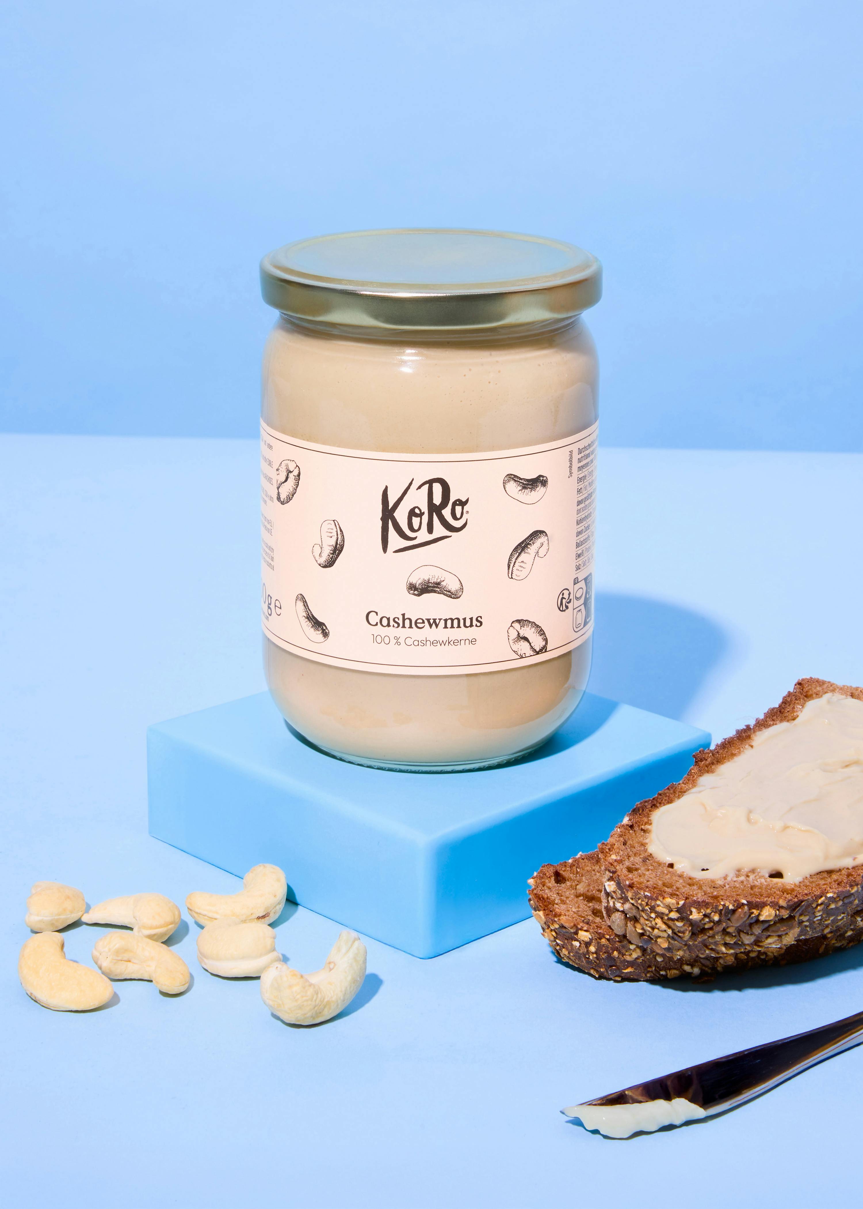 ein glas koro cashewmus steht auf einem blauen block, umgeben von cashewkernen und einem brot mit cashewmus.