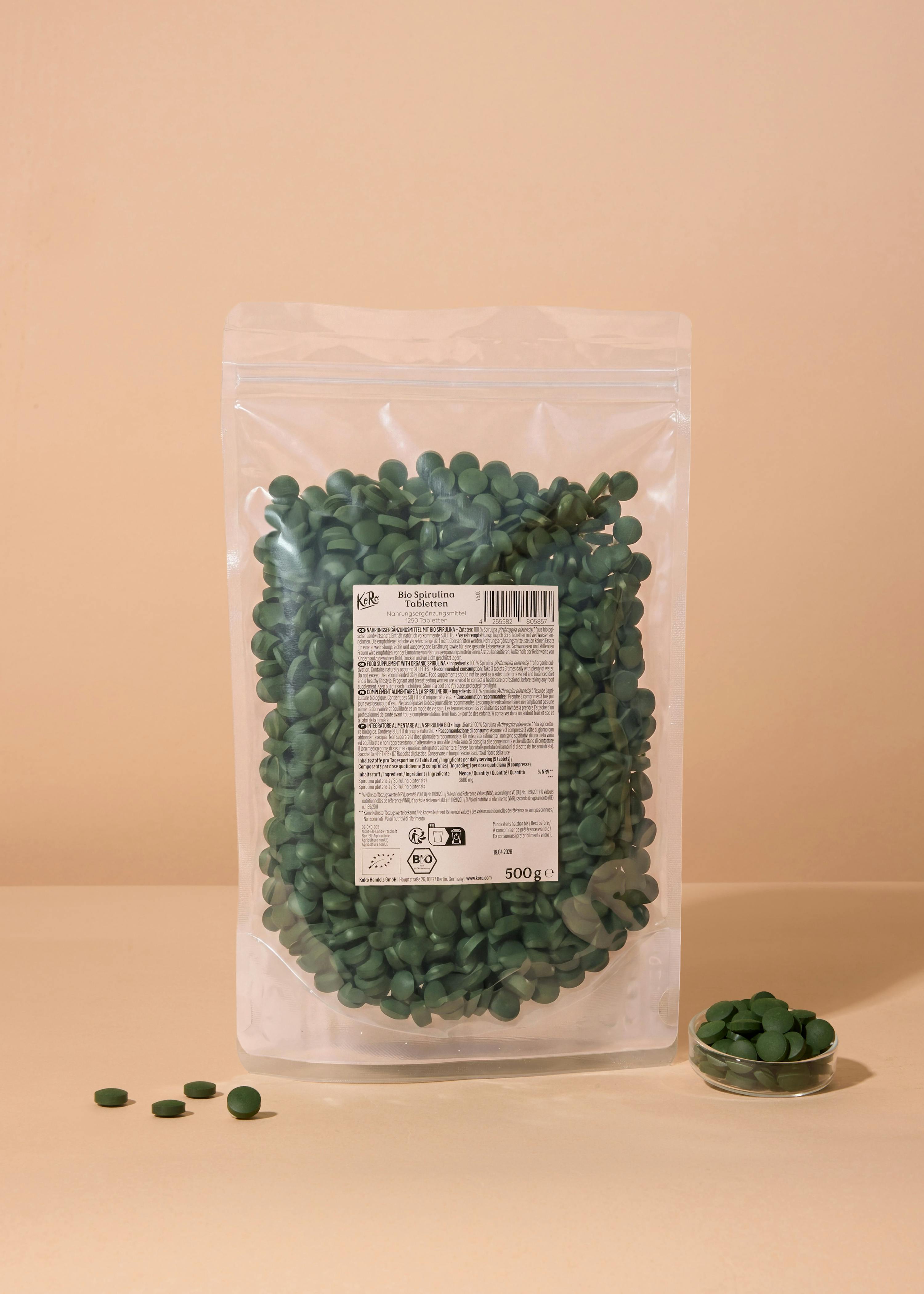eine transparente plastiktüte mit grünen bio-spirulina-tabletten steht auf einer hellen oberfläche, daneben liegen einige tabletten und eine kleine glasschale mit weiteren tabletten.