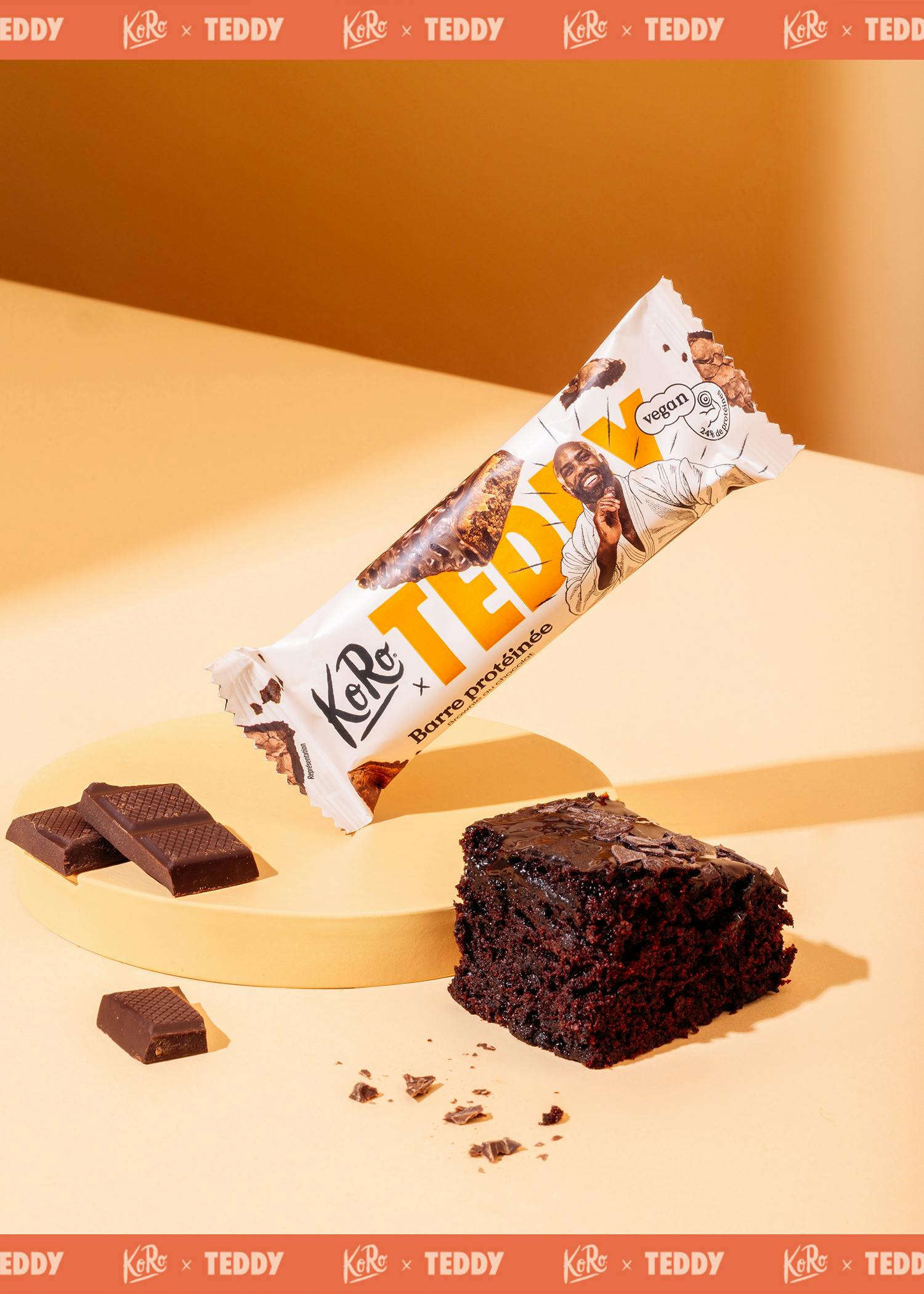 eine koro x teddy vegane schokoladen-proteinriegelpackung, schokoladenstücke und ein schokoladenbrownie auf einem gelben podest vor einem orangefarbenen hintergrund.