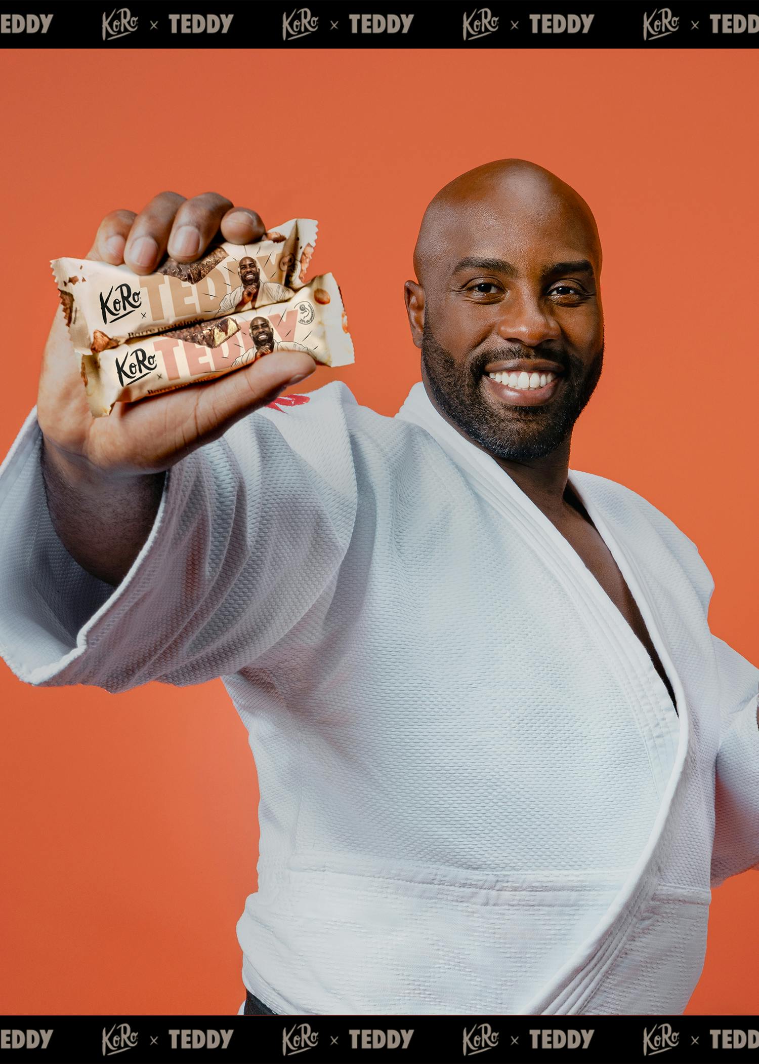 ein lächelnder mann in einem weißen judo-gi hält zwei koro x teddy snackriegel, die sein bild zeigen, vor einem orangefarbenen hintergrund.