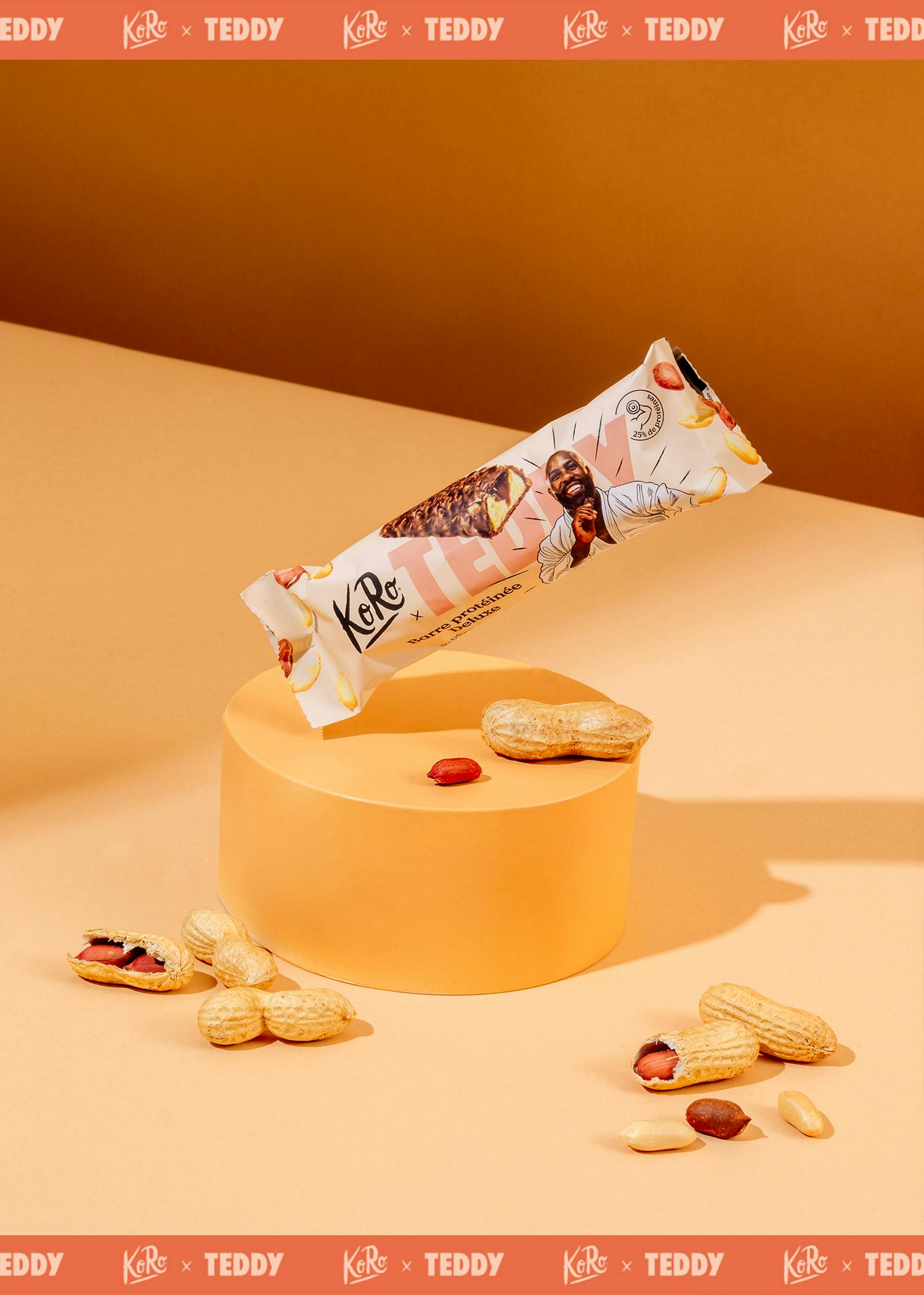 eine koro x teddy proteinriegelpackung mit einem lächelnden mann und einer aufgeschnittenen riegelansicht schwebt über einem orangefarbenen zylinder, umgeben von erdnüssen auf einem orangefarbenen hintergrund.