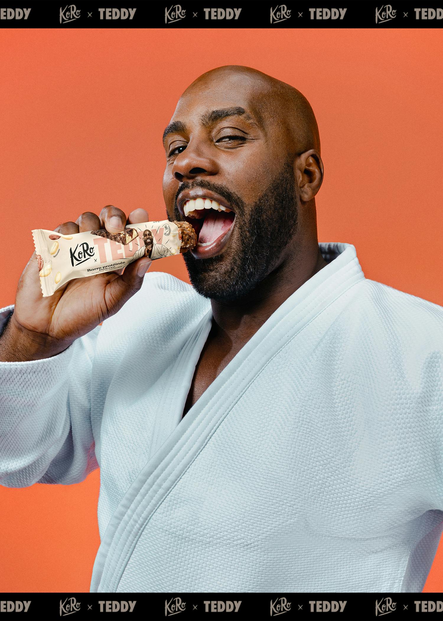 ein bärtiger mann in einem weißen gi beißt glücklich in einen koro x teddy proteinriegel vor einem orangefarbenen hintergrund.