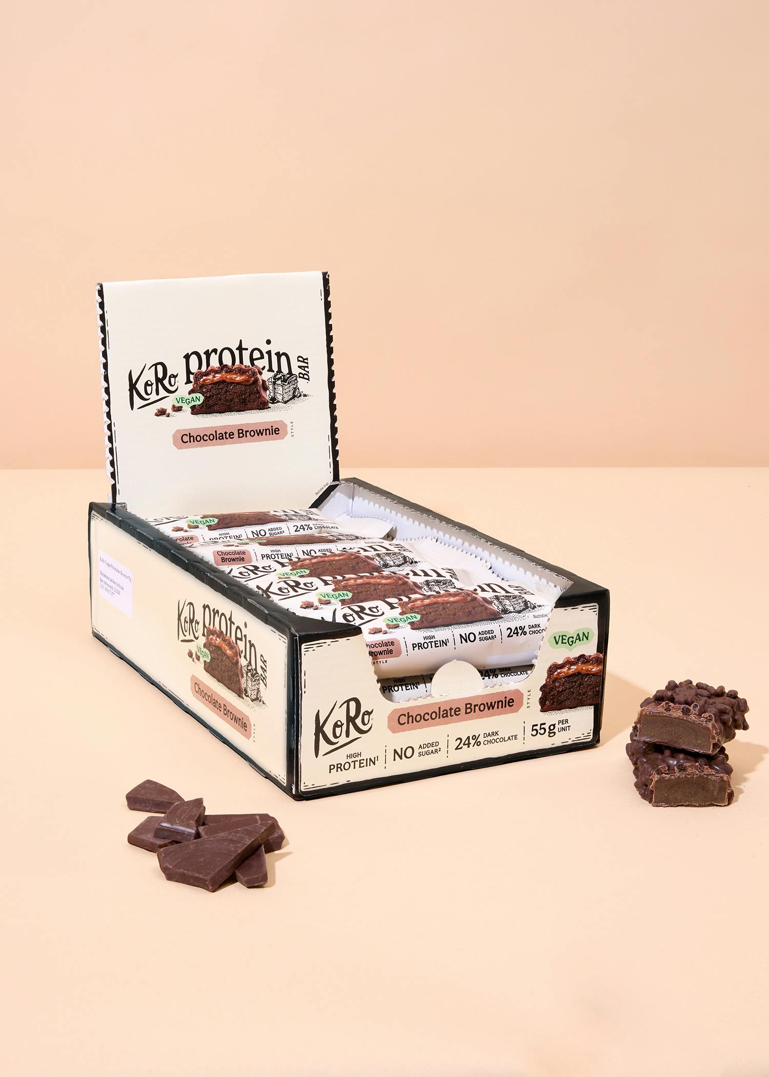 eine offene displaybox mit koro protein schoko-brownie-riegeln, die als vegan, proteinreich und ohne zuckerzusatz beworben werden, mit einigen losen schokoladenstücken und zwei angeschnittenen riegeln daneben.