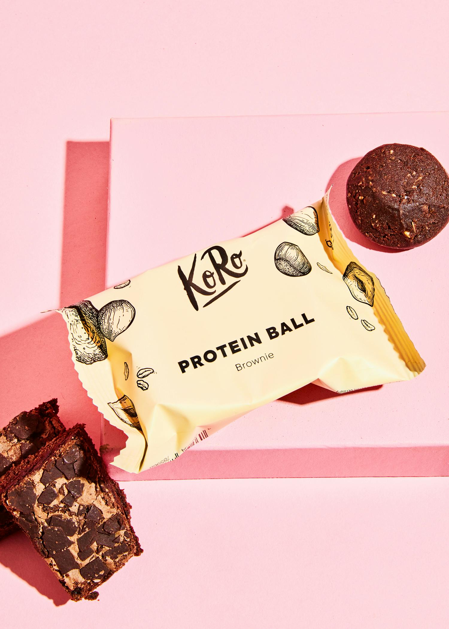 eine verpackte proteinkugel mit brownie-geschmack liegt auf einem rosa tisch neben brownies