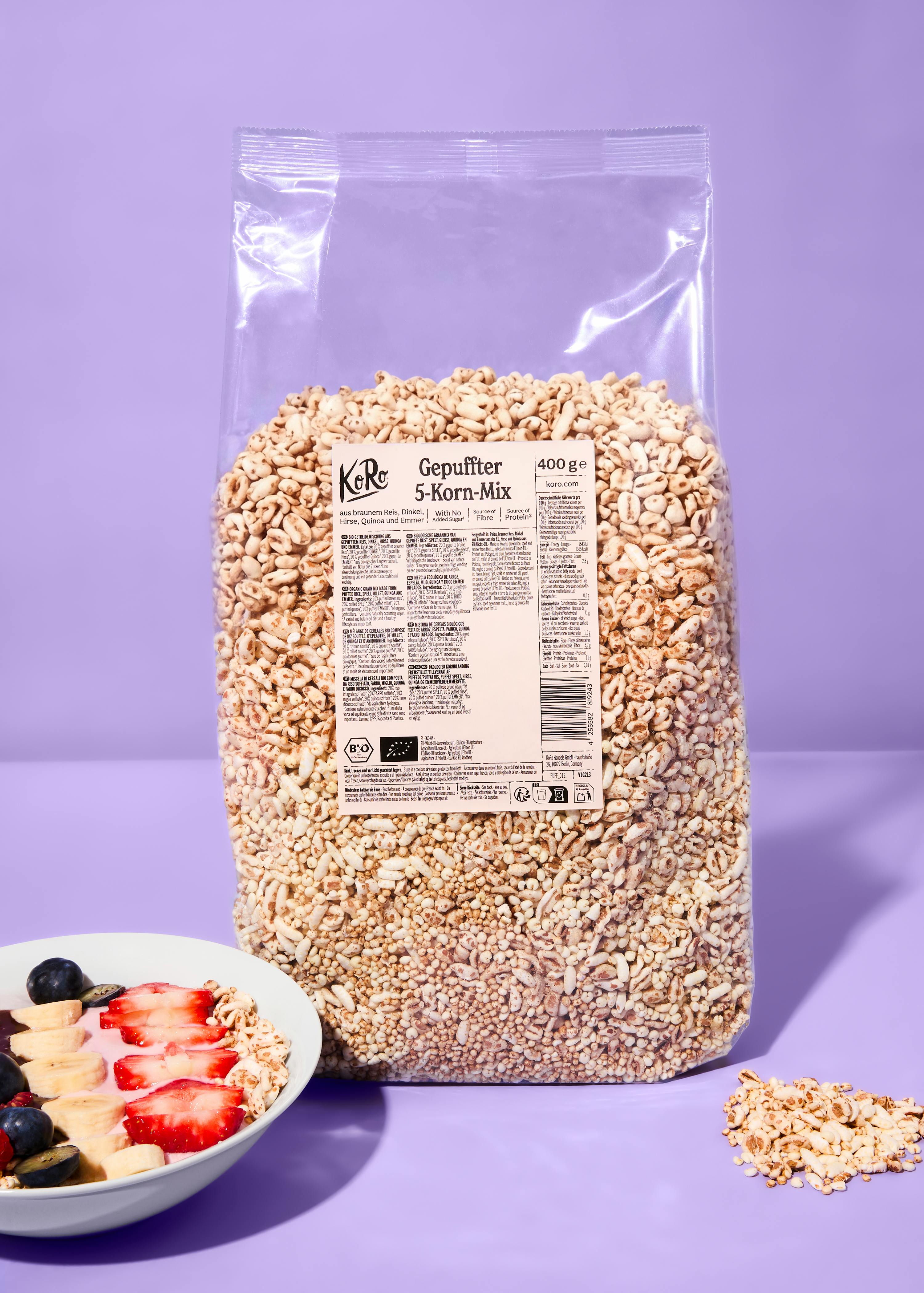 eine große, durchsichtige tüte mit gepufftem 5-korn-mix der marke koro steht neben einer schale mit joghurt, früchten und dem mix, auf einem lila hintergrund.