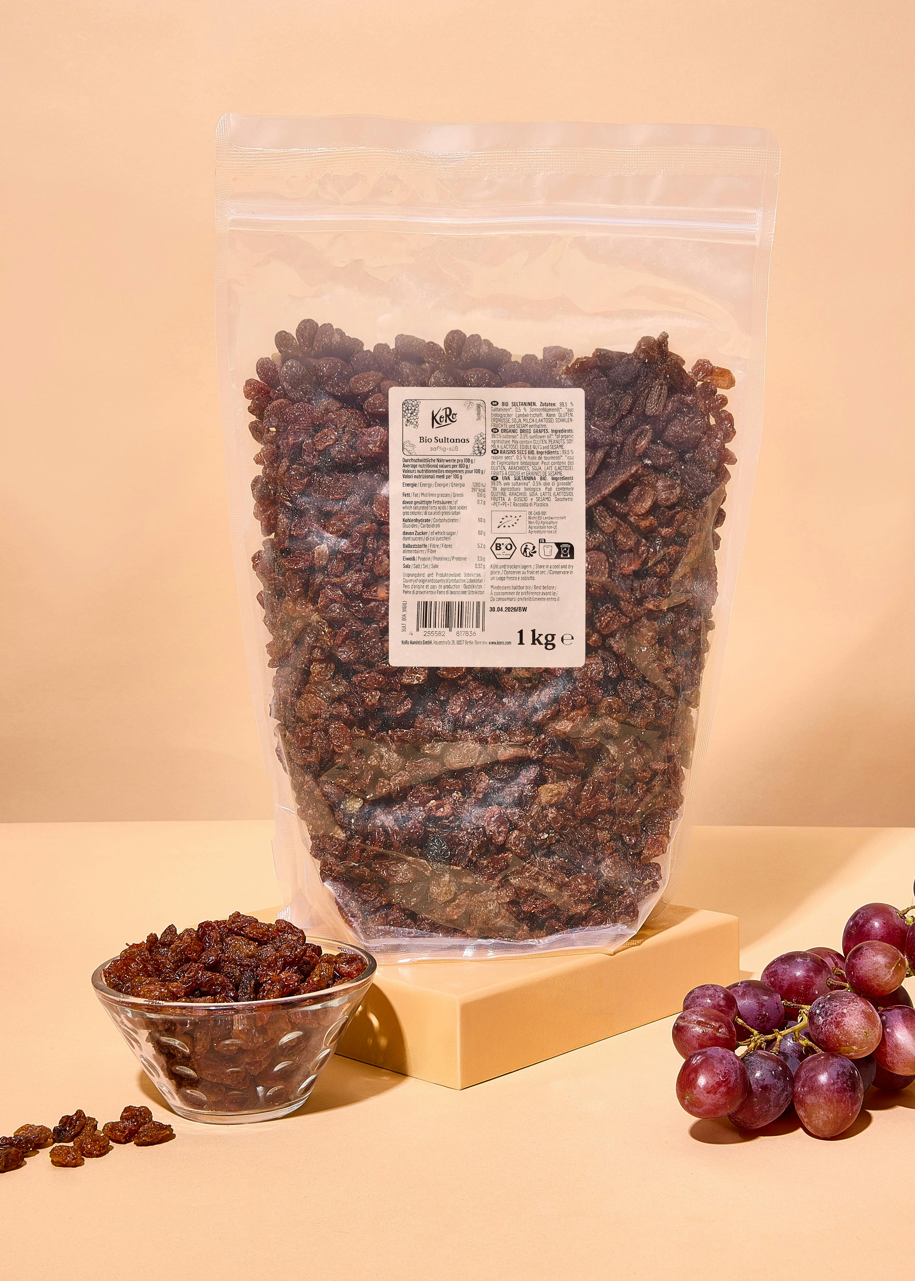 eine große, durchsichtige tüte mit 1 kg bio-sultaninen steht neben einer kleinen schale mit sultaninen und einer traube roter weintrauben auf einem hellen tisch.