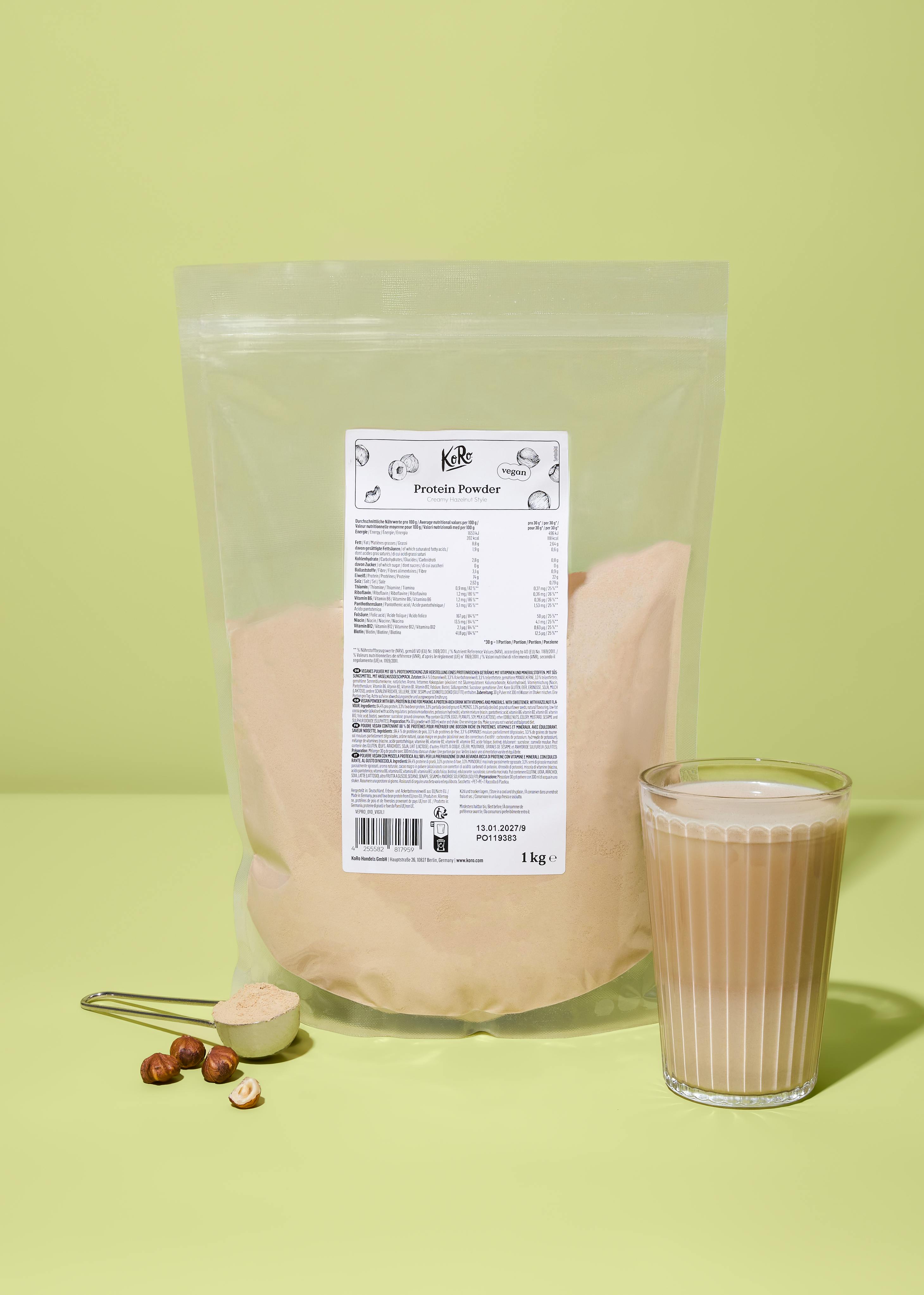 ein beutel koro proteinpulver (creamy hazelnut style, vegan, 1 kg) steht neben einem glas proteinshake, einer messschaufel mit pulver und einigen haselnüssen auf hellgrünem hintergrund.